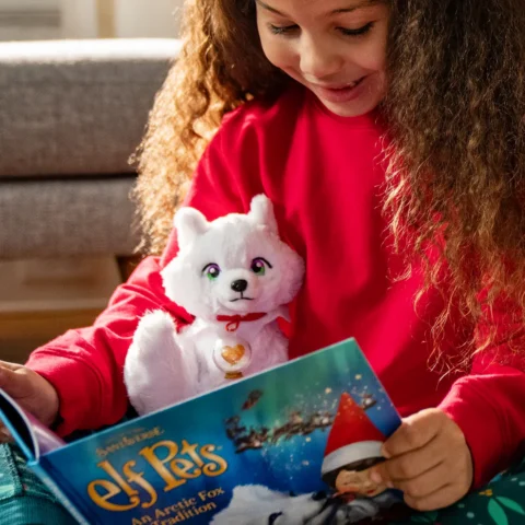 A young girl with long brown hair reads the Elf Pets arctic fox storybook while cuddling her plush arctic fox