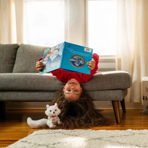 A girl wearing a red shirt hangs upside down from a sofa while reading the arctic fox storybook, as her Elf Pets arctic fox sits closeby