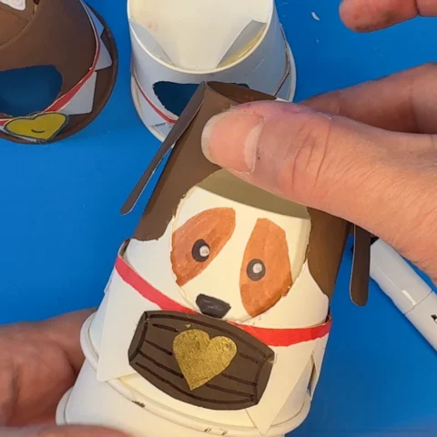 A hand holds the Elf Pets paper cup craft and places the outer cup over the inner cup