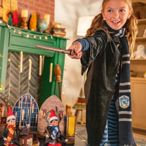 A young girl dressed in a Hogwarts robe holds a wand and pretends to cast a spell