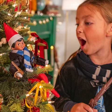 A Scout Elf holds a miniature wand as a young girl discovers the elf in a Christmas tree