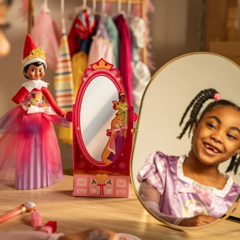A young black girl with beautiful braids smiles into a mirror beside her Elf on the Shelf which is standing beside a toy princess mirror