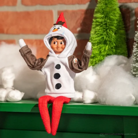 The Elf on the Shelf, wearing an Olaf hoodie, sits on a shelf with his arms raised in celebration
