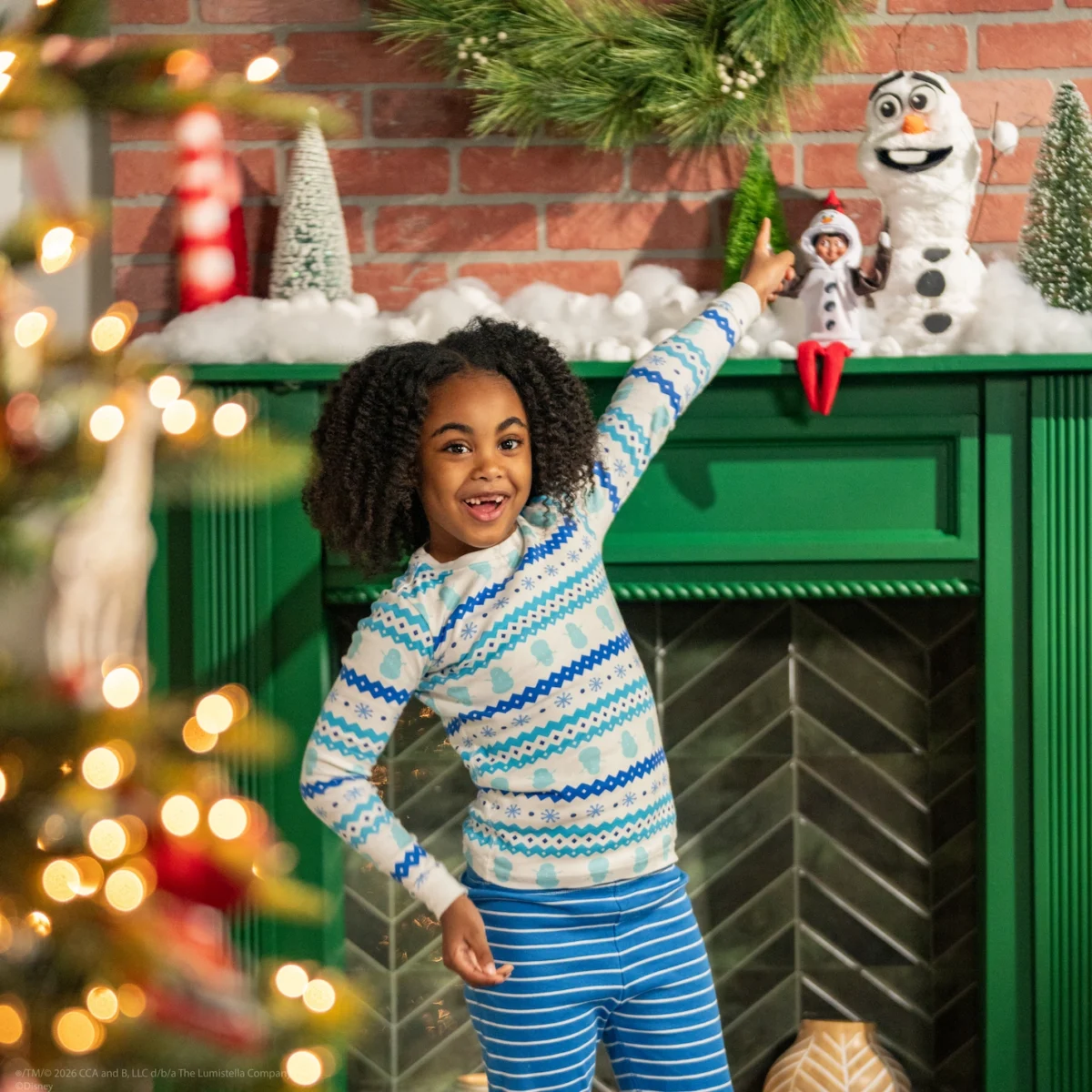 A young girl points to her Elf on the Shelf and the snowy scene he has created with Olaf above the fireplace