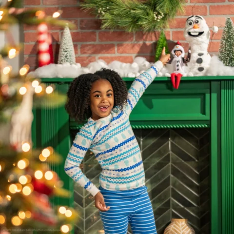 A young girl points to her Elf on the Shelf and the snowy scene he has created with Olaf above the fireplace
