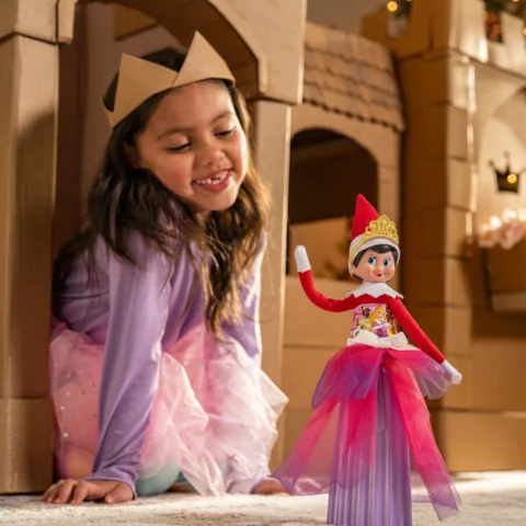 A young girl wearing a cardboard crown sits in the entrance to a play castle made from cardboard while smiling at her Elf on the Shelf dressed in a princess costume