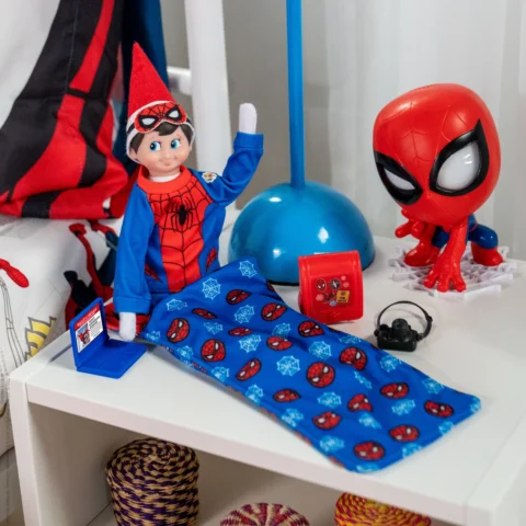 The Elf on the Shelf Scout Elf, dressed in Spider-man pajamas, sits in a Spider-man-themed sleeping bag with accessories