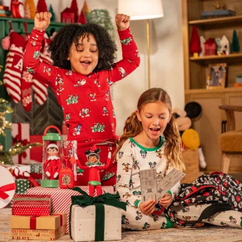 A young boy and girl hold travel tickets as their Elf on the Shelf sits atop a present with a toy suitcase showing he plans to travel along with them