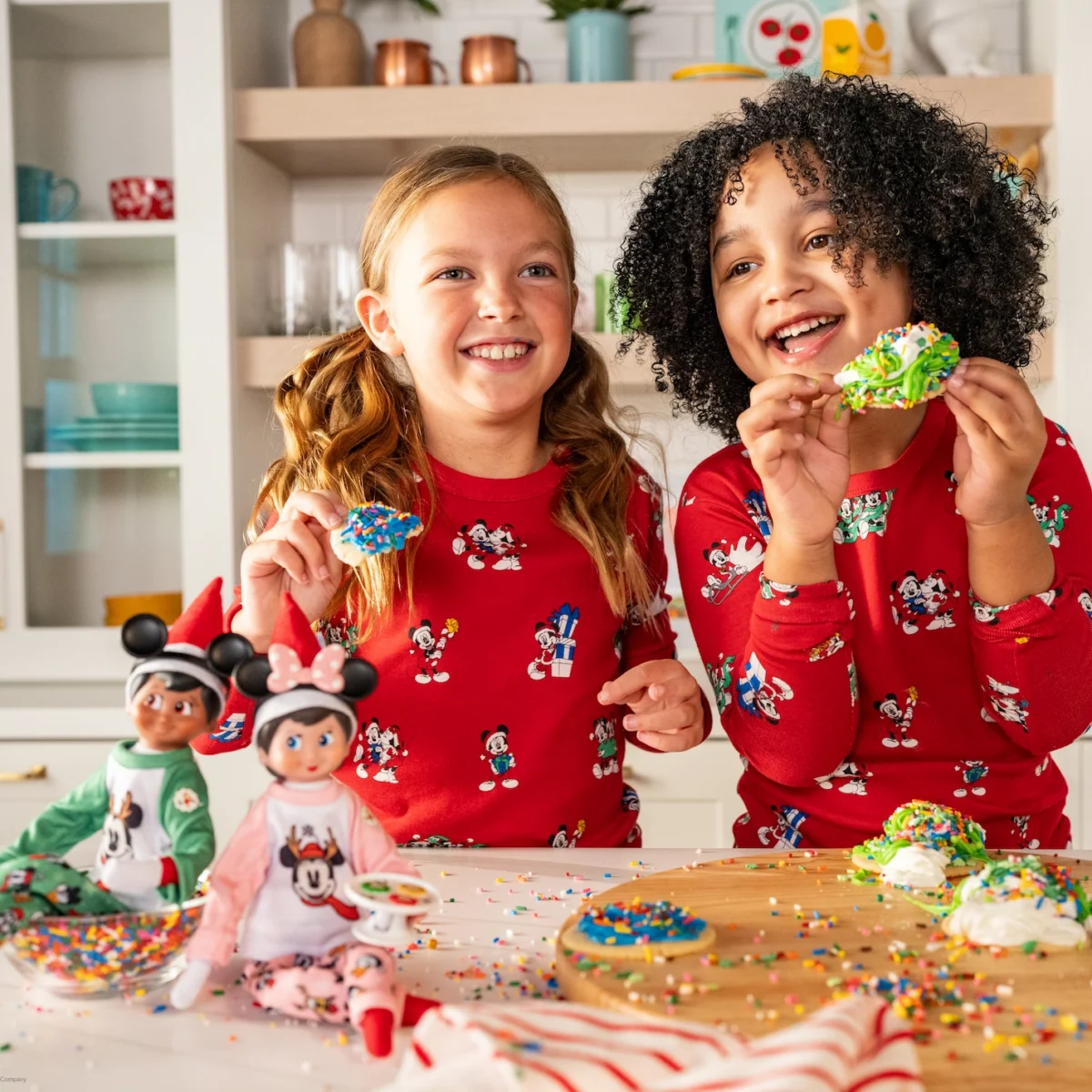 A boy and girl decorate cookies alongside their Scout Elf Girl and Scout Elf boy who are dressed in Mickey and Minnie pajamas
