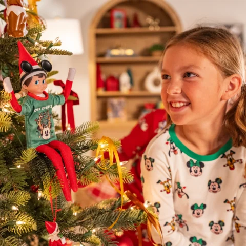 A young blonde girl smiles at The Elf on the Shelf who is wearing a Disney Mickey & Friends Oh Deer Tshirt