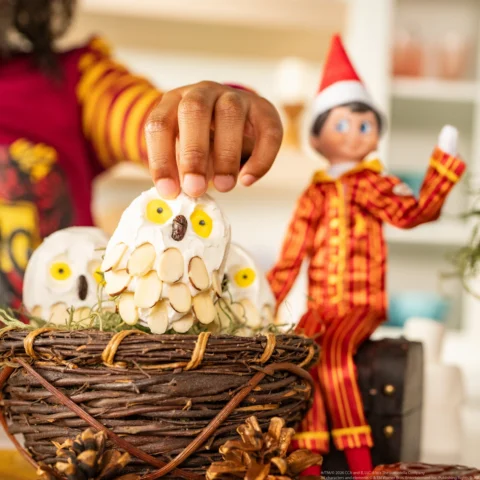 A Scout Elf in Harry Potter pajamas sits beside a basket of Potter-themed owls as a young girl's hand picks up one of the owls
