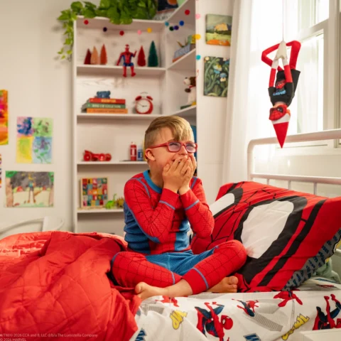 A young boy sits in his Spider-man themed bedroom and covers his mouth, laughing as he watches his Scout Elf hanging upside down dressed in a Spider-man tshirt