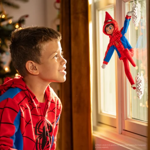 A young boy looks at his Elf on the Shelf dressed in a Spider-man hoodie as the elf clings to snowflake shaped spider suction cups