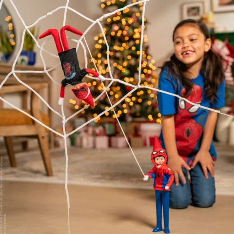 A little girl in a Spider-man tshirt watches as an Elf on the Shelf Scout Elf pretends to sling a web and catches another elf in it
