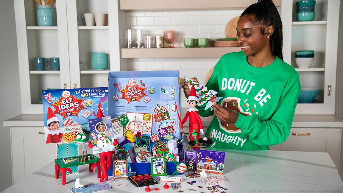 A woman standing in a kitchen behind a counter. The Elf Ideas Fun and Games Kit is sitting on the counter and the box is open and all the contents are set up around it. She is smiling while holding the cut out Christmas tree prop.
