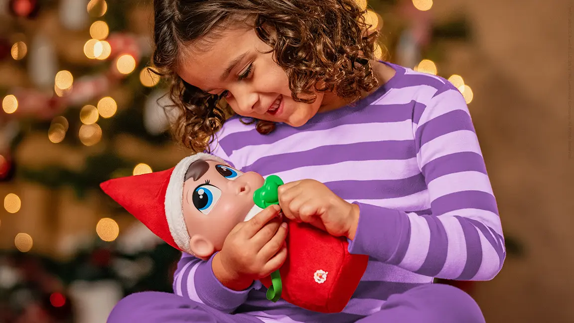 A girl cradling a Frost Pips plush and smiling down at him. She is holding the pacifier close to his mouth.