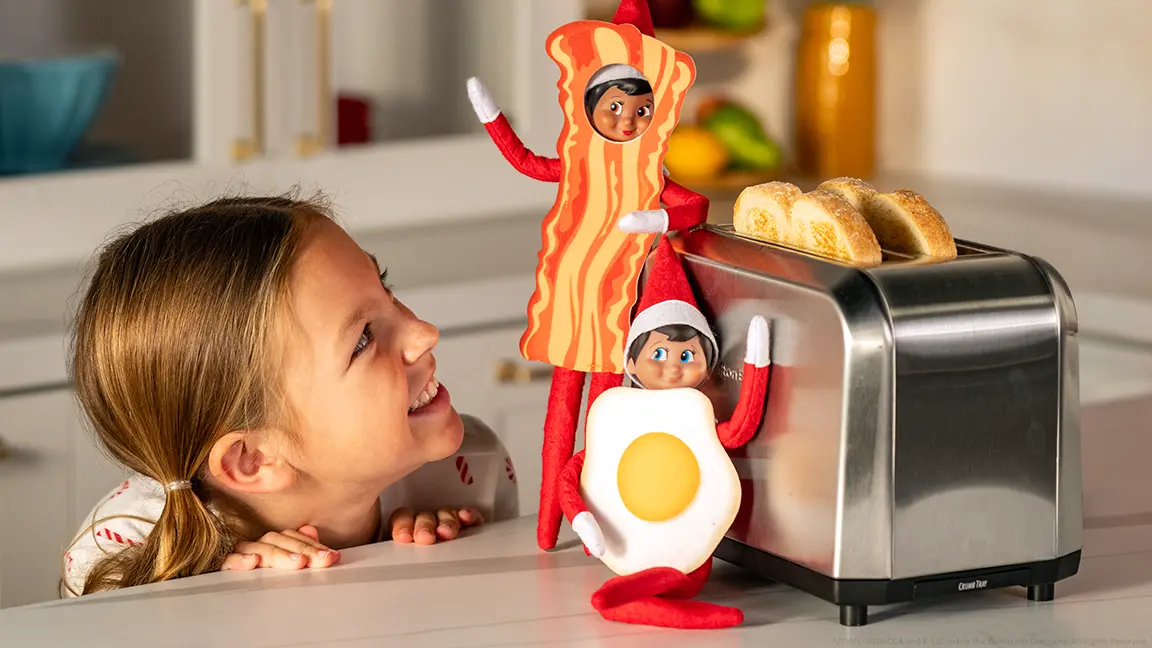 Two Scout Elves wearing the Bacon and Eggs Bestie Bundle Costume Set next to a toaster sitting on a kitchen countertop. A girl is peeking up from behind the counter looking at the elves and smiling.