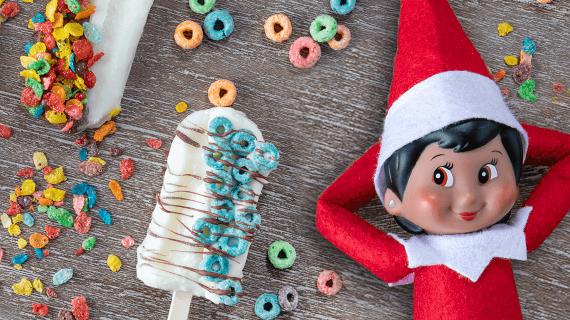 A girl Scout Elf leaning back on a wooden surface with her hands behind her head. Next to her are popsicles decorated with various cereal pieces.