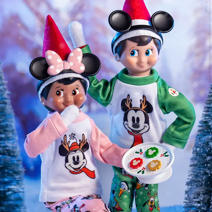 Boy and Girl Scout Elves wearing the Disney's Mickey & Friends Cookies for Santa PJs set. Behind them is a winter scene with snow-covered pine trees.