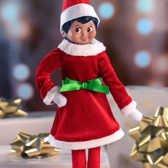A girl Scout Elf wearing the Sleighing as Santa Party Dress. She is standing on a gold and white Christmas gift, and more gifts are stacked behind her. In the foreground are elf-sized snow-covered pine trees.