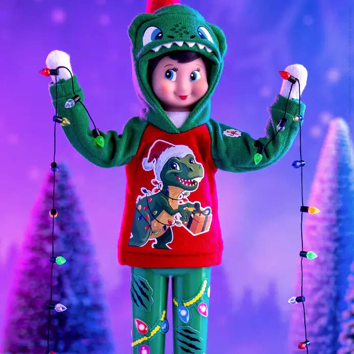 A girl Scout Elf wearing the MagiFreez® Dashing Dino Elf Hoodie & Standing Pants. She is in a snowy, winter scene and is holding up a string of elf-sized Christmas lights. Multiple Christmas gifts are sitting on the ground.
