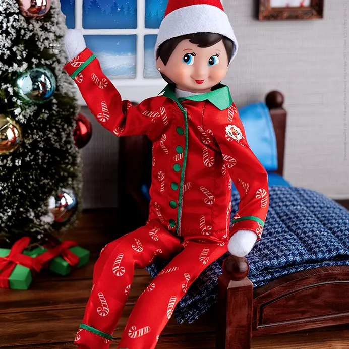 A girl Scout Elf sitting on an elf-sized bed wearing the Candy Cane Couture pajama set. Behind her is a little Christmas tree, a picture frame on the wall, and a window with a snowy, winter scene outside.