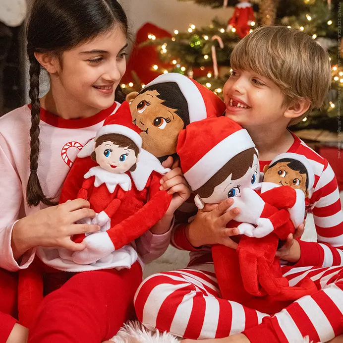 A girl and a boy sitting on the floor in front of a Christmas tree. They are holding several different sized Plushee Pals and smiling at each other.
