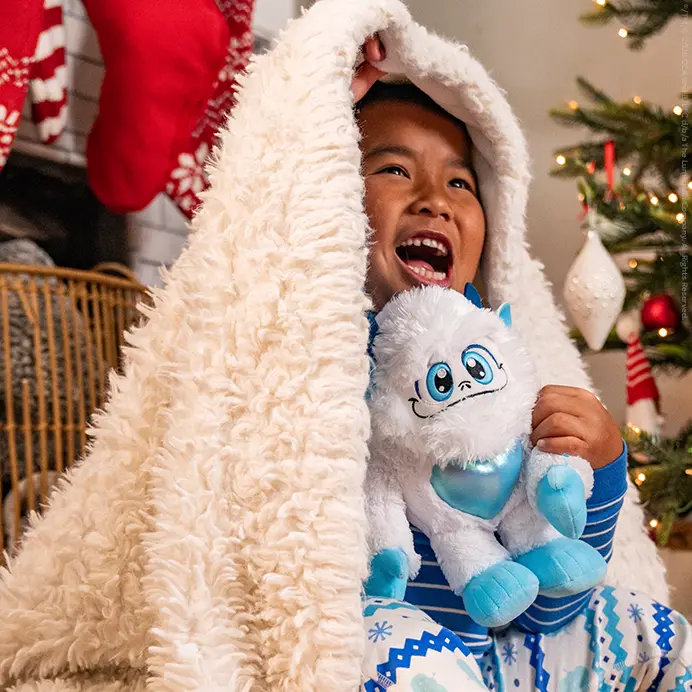 A boy sitting on the floor holding the Little Grunt plush. He is peeking out from underneath a fuzzy white blanket. Behind him are stockings hung on a mantel and a Christmas tree.