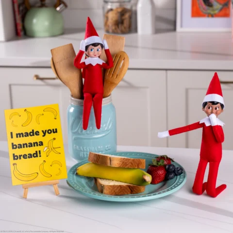 Two Scout Elves playfully point to a an unpeeled banana between two slices of bread as their banana bread recipe