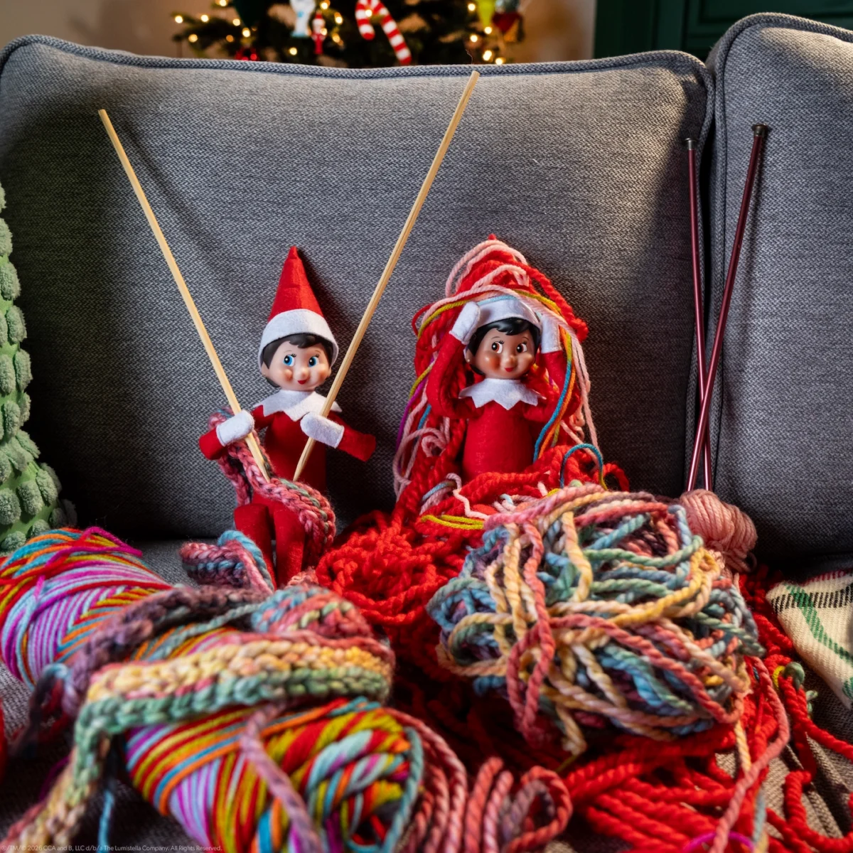 Two Scout Elves sit in a tangle of yarn, appearing to try to knit a scarf