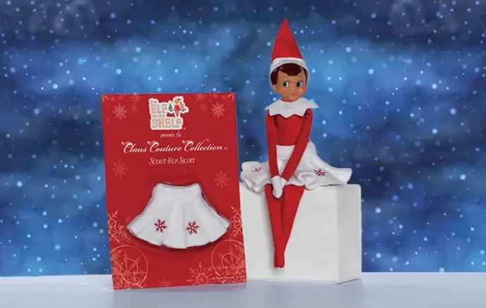 A girl Scout Elf dressed in the first Claus Couture Collection piece, the Snowflake Skirt, and sitting next to the Snowflake Skirt in its packaging