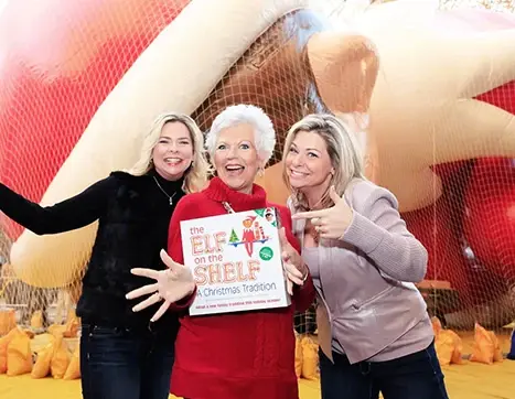 Christa, Carol, and Chanda holding a The Elf on the Shelf boxset in front of the Macy's Thanksgiving Day Parade Scout Elf balloon