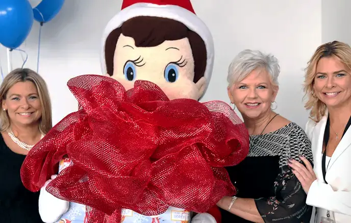 Scout Elf mascot holding a gift with a big bow standing in between Chanda, Carol, and Christa