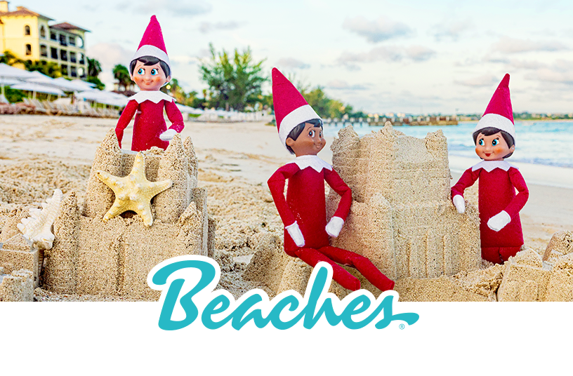 Three Scout Elves sitting around 2 sandcastles on a beach. There is a starfish and a shell sitting on one of the sandcastles. The shore, a line of beach chairs and umbrellas, a waterfront property and a tree line are in the background. The Beaches logo overlays the bottom center of the photo.