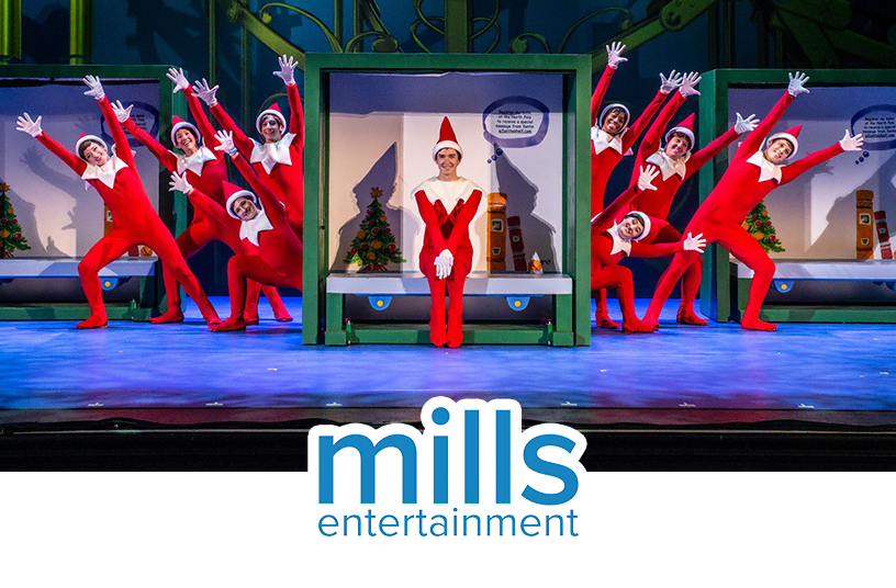 Actors dressed in Scout Elf outfits dancing on a stage during the The Elf on the Shelf musical production. In the middle of the stage there is a large re-creation of the inside of The Elf on the Shelf boxset and there is an actor dressed in a Scout Elf costume sitting on the shelf. The Mills Entertainment logo overlays the bottom center of the photo.