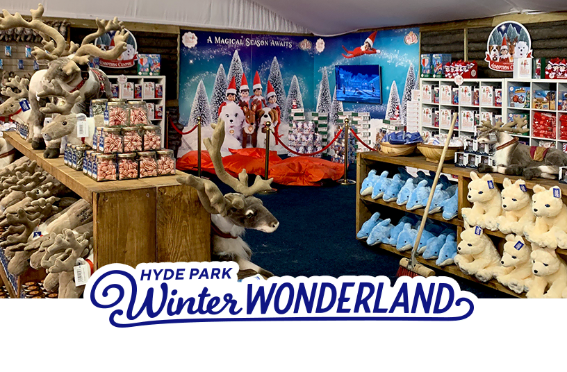 The Hyde Park Winter Wonderland gift shop with a display in the corner containing stacks of The Elf on the Shelf and Elf Pets boxsets. The area is partially surrounded by a red velvet rope and there are red satin pillows on the ground. There is a backdrop containing photos of The Elf on the Shelf Dolls, Elf Pets plushes, a blue and teal sky with white sparkle trails. The Elf on the Shelf and Elf Pets logo are at the top of the backdrop. Along the wall on the right are white cube shelves containing The Elf on the Shelf boxsets, Scout Elves at Play products, Plushee Pal clip-ons and clothing. Above the shelf is a sign that says Adoption Center. The Hyde Park Winter Wonderland logo overlays the bottom center of the photo.