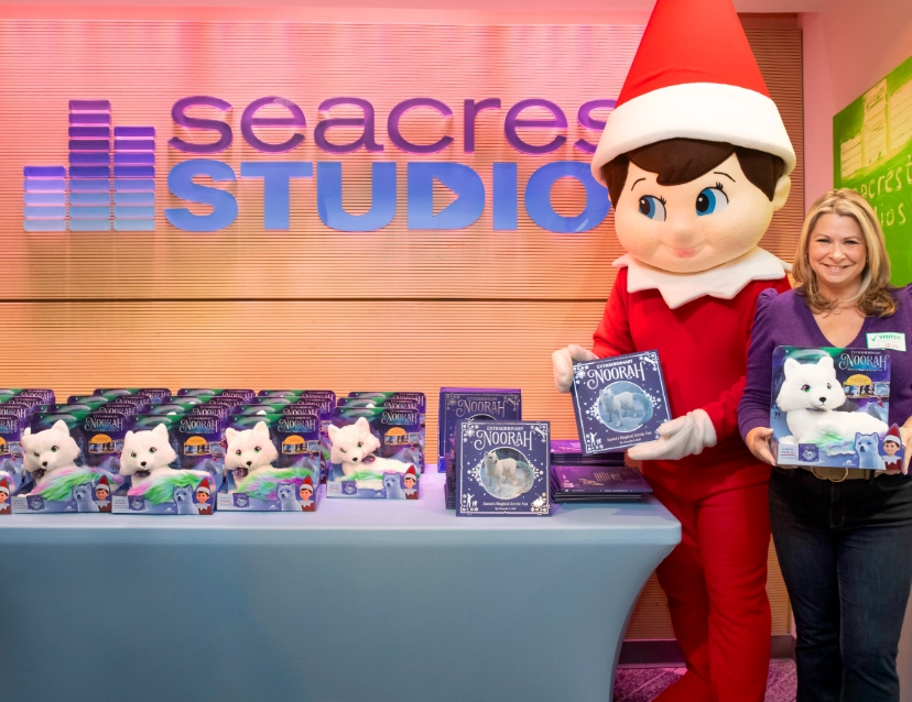 Chanda Bell holding an Extraordinary Noorah Plush in its packaging standing next to a Scout Elf Mascot holding the Extraordinary Noorah storybook. They are standing next to a table with rows of Extraordinary Noorah plushes and stacks of Extraordinary Noorah storybooks. There is a sign on the wall above the table that reads Seacrest Studios.