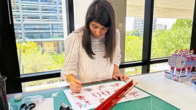 A woman leaned over a desk working on a licensing product project. On the desk is a printout, a ruler, tape, scissors, and a cutting mat. Behind the woman is a window overlooking trees and office buildings.