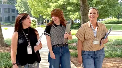 Three women smiling and laughing as they walk outside of the office building. In the background are trees, grass, a sidewalk and pinestraw