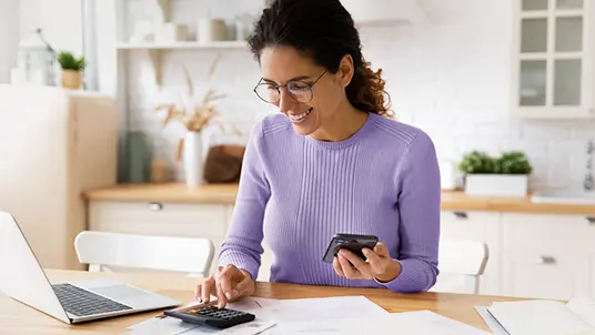 A woman using a calculator sitting at a kitchen table. On the table is an open laptop, as well as scattered paperwork. A kitchen scene is behind the woman.