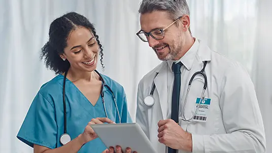 Two medical professionals looking at a tablet while smiling. The female on the left is in blue scrubs, and the male on the right is wearing a white coat and blue tie. They both have stethoscopes around their necks. White, sheer curtains are in the background.