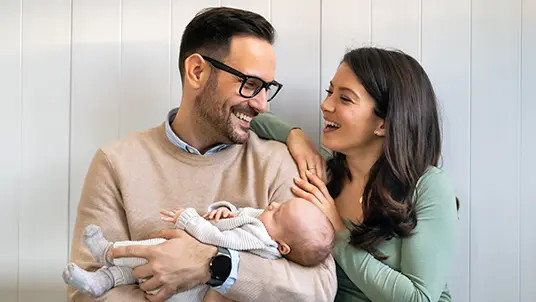 A smiling father holding an infant in his arms. The mother has her arms draped around the fathers shoulder. The parents are smiling while they look at each other. A white wall is in the background.