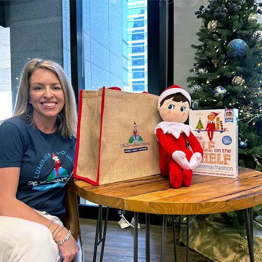 A woman in a Lumineer Volunteer shirt sitting next to a table. On the table is a canvas bag with The Lumistella Company philanthropy logo, a Plushee Pal boy and a The Elf on the Shelf boxset. In the background is a Christmas tree and a window.