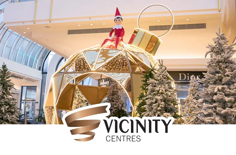 A large Scout Elf figure sitting on top of a large, geometric, hollow Christmas ornament sculpture that is part of Christmas activation in a Vicinity Centre mall. The space in and around the sculpture is filled with flocked pine trees. The Dior store is right behind the activation.