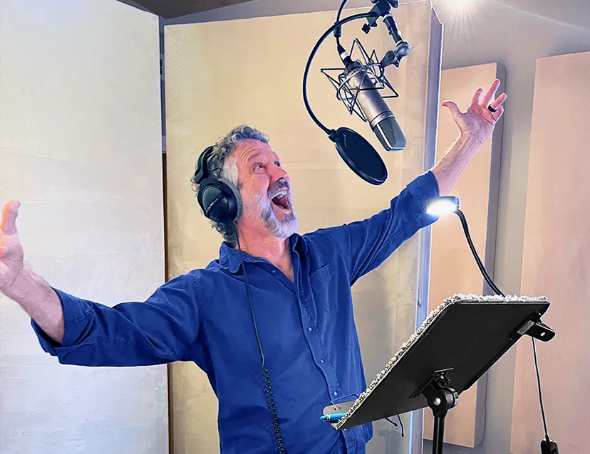 A man in a recording booth gesturing grandly with both arms open as he speaks into a microphone.