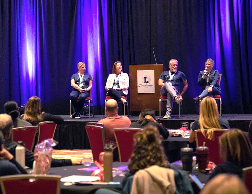 Executive team sitting on a stage in front of a podium and curtains in a conference room at a Lumiversity event. Chanda Bell has a microphone held to her mouth. Employees are sitting at round tables on the main floor.
