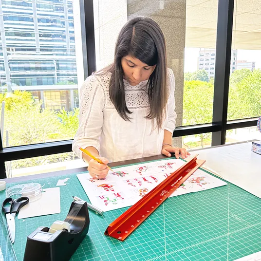 A woman leaned over a desk working on a licensing product project. On the desk is a printout, a ruler, tape, scissors, and a cutting mat. Behind the woman is a window overlooking trees and office buildings.