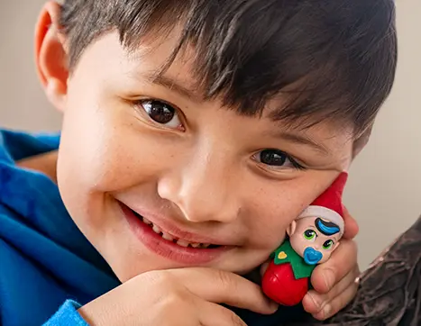 Close up of a boy's face who has brown hair and brown eyes. He's holding a Frost Pip up to his cheek and smiling. The Frost Pip has a blue streak in his hair, a blue pacifier in his mouth, and green eyes.