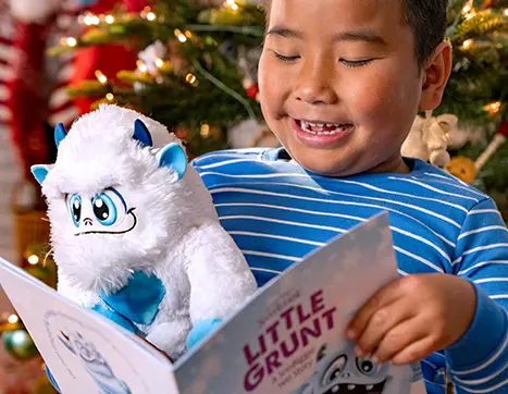 A boy with black hair and a blue and white stripe shirt from the chest up. He is looking down at an open Little Grunt story book. In the crook of his arm is a Little Grunt plush. In the background is a Christmas tree.