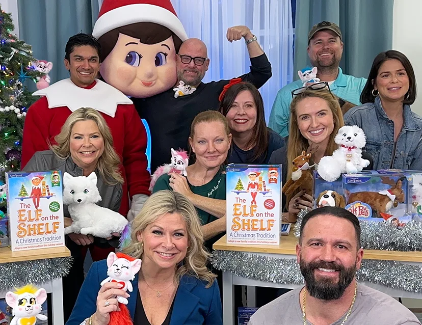 The Lumistella Company employees huddled together holding various plush products. In between them are 2 tables with The Elf on the Shelf and Elf Pets boxsets on them. The tables are decorated with silver garland. One of the employees is wearing an elf mascot suit and another employee is holding the head of the mascot suit on his shoulder. In the background is a Christmas tree, white walls and green and white, sheer curtains.